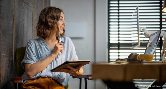 Junge Frau sitzt mit Tablet vor einem Tisch, auf dem ein Laptop steht.