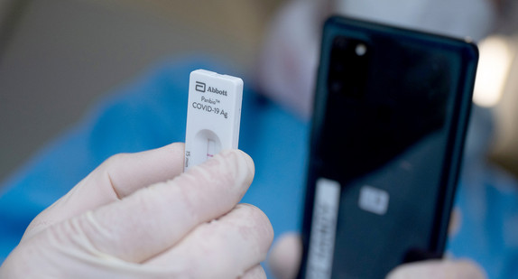 Hand in Schutzhandschuh hält einen Corona-Schnelltest vor ein Smartphone