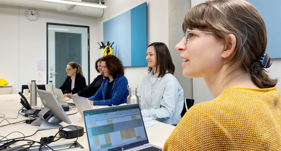 Junge Frau sitzt mit vier anderen Frauen in einem Konferenzraum, auf dem Tisch stehen mehrere Laptops.