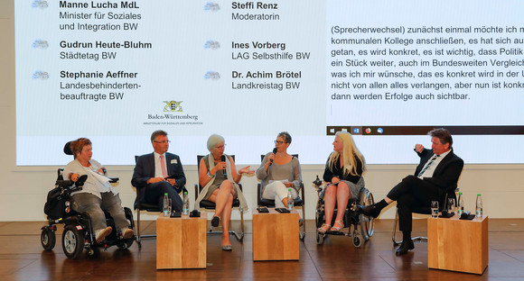 Podiumsdiskussion auf der Bühne mit Ines Vorberg (Mitglied des Vorstands der LAG Selbsthilfe Baden-Württemberg e. V.), Landrat Dr. Achim Brötel (Vizepräsident des Landkreistags Baden-Württemberg), Oberbürgermeisterin a. D. Gudrun Heute-Bluhm (Vorstandsmitglied Städtetag Baden-Württemberg), Moderatorin Steffi Renz, Landes-Behindertenbeauftragte Stephanie Aeffner und Minister Manne Lucha