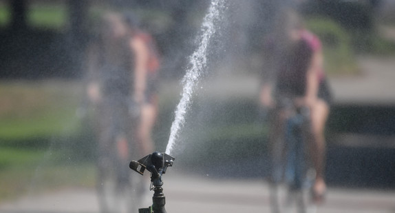 Junge Menschen fahren auf Fahrrädern an einem Wassersprenger vorbei.