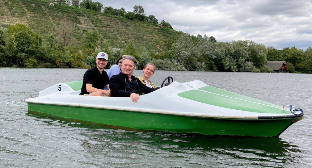 Minister Lucha sitzt mit drei baden-württembergischen Jugendgemeinderäten in einem grün-weißen Tretboot. Zusammen fahren sie über den Max-Eyth-See. Im Hintergrund befindet sich ein Hang mit Weinreben.