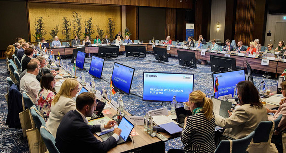 Teilnehmende der Jugend- und Familienministerkonferenz 2026 sitzen mit Blick auf mehrere kreisförmig angeordnete Bildschirme an einem großen Konferenztisch.