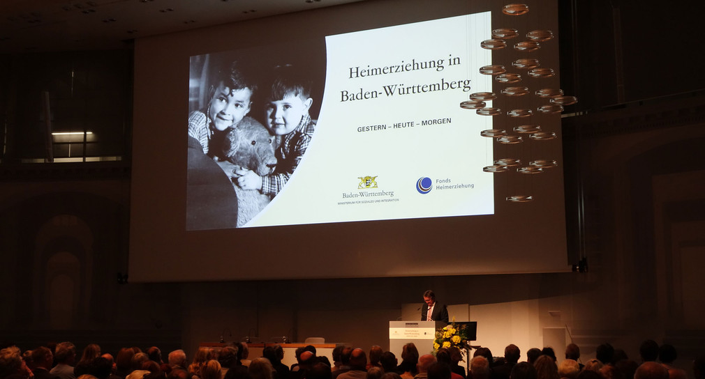 Sozialminister Manne Lucha spricht auf der Bühne in Veranstaltungssaal (Bild: © Ministerium für Soziales und Integration Baden-Württemberg)