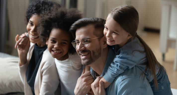 Auf einer Couch sitzt eine Familie mit zwei Kindern. Eines der Kinder hat dunklere Haut und krauses Haar wie die Mutter. Alle lachen und scheinen das Zusammensein zu genießen.