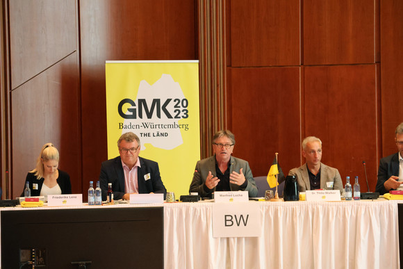 Minister Manne Lucha sitzt mit anderen Teilnehmenden in Konferenzsaal, im Hintergrund eine Stellwand mit GMK-Logo