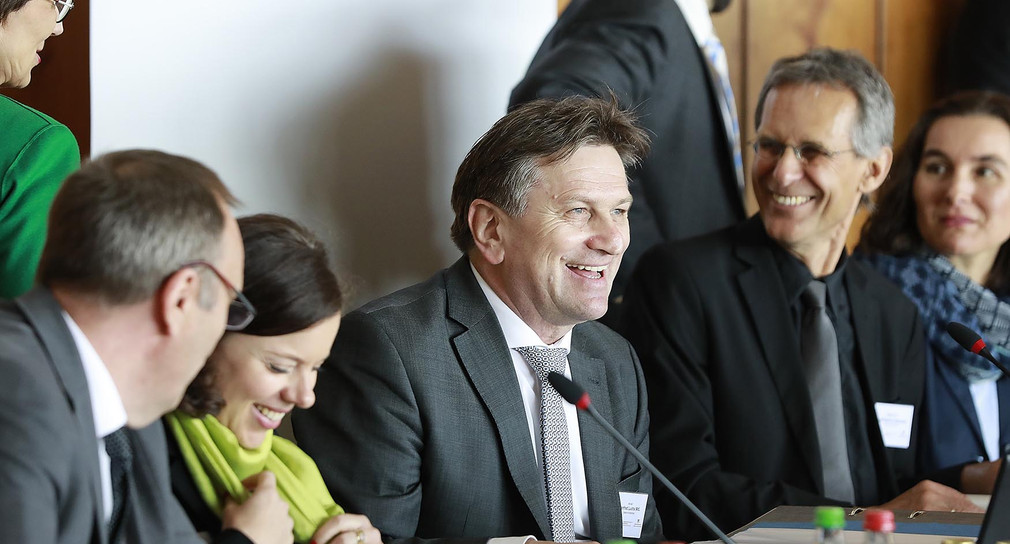 Minister Manne Lucha sitzt umringt von anderen Teilnehmern lachend an Konferenztisch