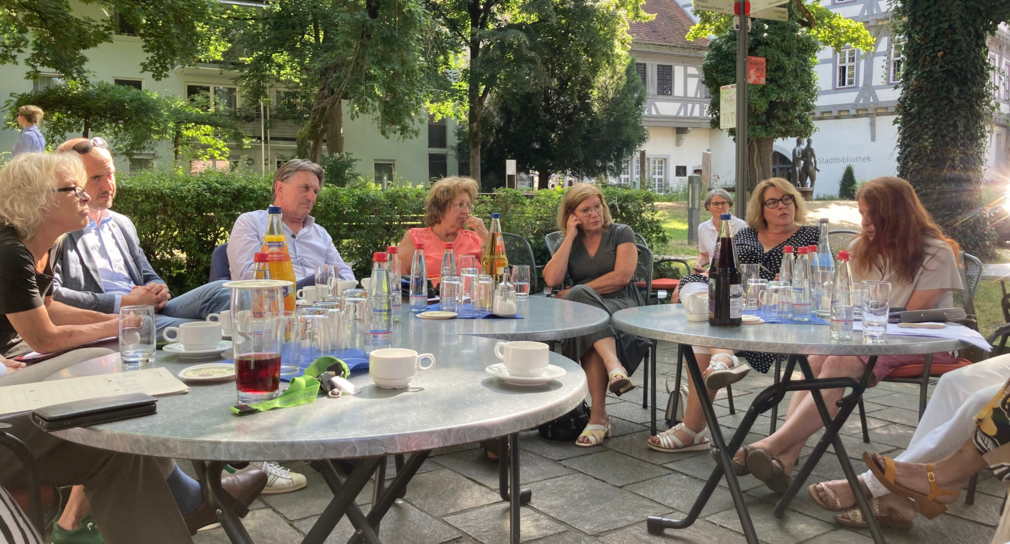 Minister Manne Lucha im Gespräch mit Mitgliedern der Aktion „Gmünd isst besser - gemeinsam gesund genießen“. Die Gesprächsteilnehmenden sitzen in einem schattigen Biergarten.