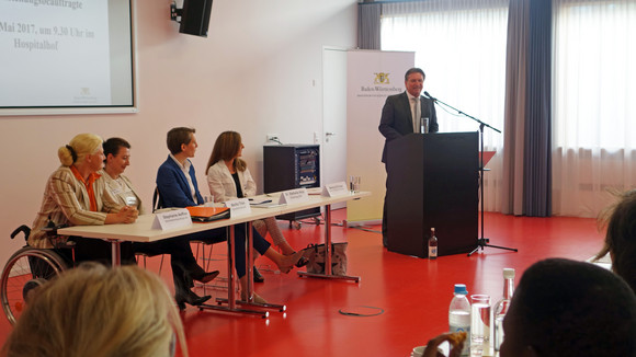 Landes-Behindertenbeauftragte Stephanie Aeffner, Melitta Thies (Sprecherin der LAG), Dr. Stefanie Hinz (Städtetag Baden-Württemberg) und Genny Di Croce (Referatsleiterin im Sozial- und Integrationsministerium) hören Minister Manne Lucha bei seiner Begrüßung der Veranstaltungsteilnehmenden zu.