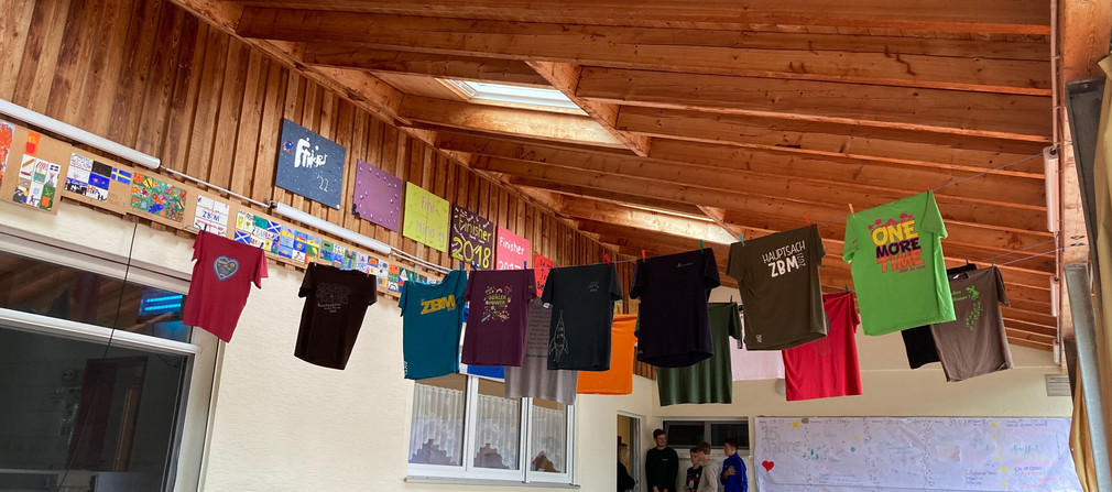 Unter dem Dach im Eingangsbereich der Zimmerbergsmühle des Kreisjugendrings Ostalb hängen viele bunte T-Shirts. 