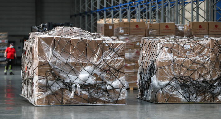 Mitarbeiter verladen in einer Lagerhalle medizinische Schutzkleidung in Form von Schutzanzügen und Masken.