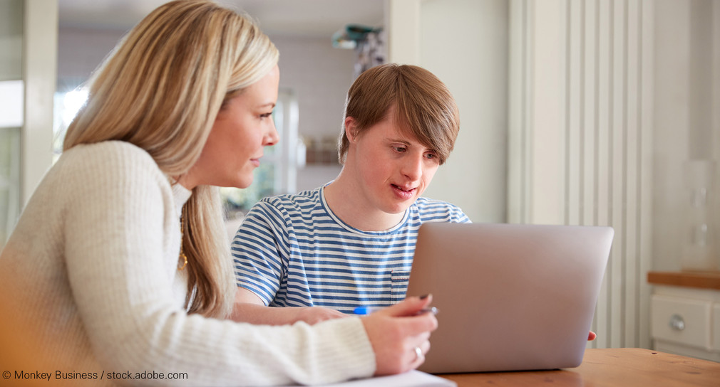 Frau sitzt mit jungem Mann mit Down-Syndrom vor einem Laptop