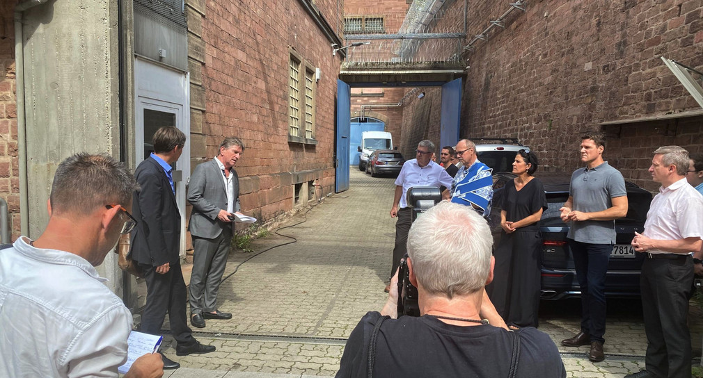 Minister Manne Lucha steht gemeinsam mit Verantwortlichen der Stadt Heidelberg in der Zufahrt zum Maßregelvollzug Fauler Pelz in Heidelberg. Fotografiert wird er dabei von Medienvertretern.
