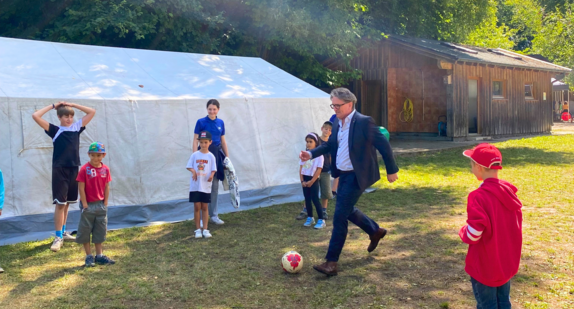 Minister Manne Lucha zeigt auf der Wiese sein Fußballkönnen. Rechts und links von ihm stehen Kinder, die das Ganze kritisch beobachten.