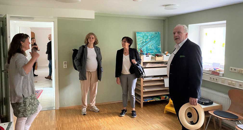 Staatssekretärin Dr. Ute Leidig steht mit Verantwortlichen des Mehrgenerationenhauses in einem Zimmer. Neben ihr steht ein Real voller Leinwände und Farben. Auf dem Real ein Aquarell in Grün und Blau.