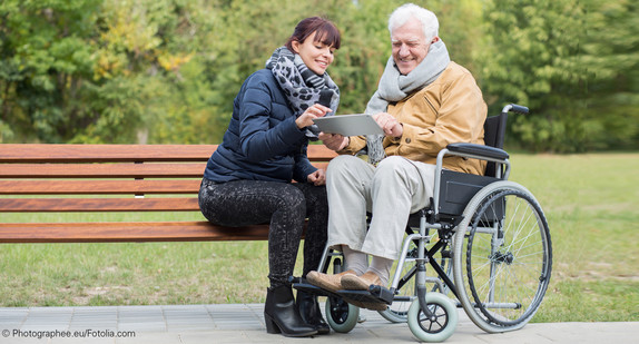 Junge Frau auf Parkbank und älterer Mann in Rollstuhl schauen auf ein Tablet