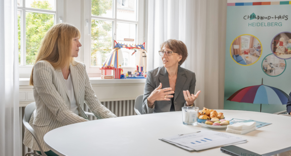Staatssekretärin Dr. Ute Leidig lässt sich von einer Verantwortlichen des Childhood-Hauses am Universitätsklinikum Heidelberg ihre Arbeit erklären. Beide sitzen dabei an einem weißen Tisch. Auf der Fensterbank im Hintergrund steht Kinderspielzeug. Rechts ein buntes Plakat, das über die Funktion des Hauses informiert. Die Farben sind sehr hell und freundlich.