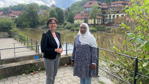 Staatssekretärin Dr. Ute Leidig und Frau Hala Elamin, die interkulturellen Promotorin des Vereins Freundeskreis Afrika e. V. stehen für ein Gruppenbild auf der Terrasse. Im Hintergrund sieht man ein idyllisches Flusspanorama.