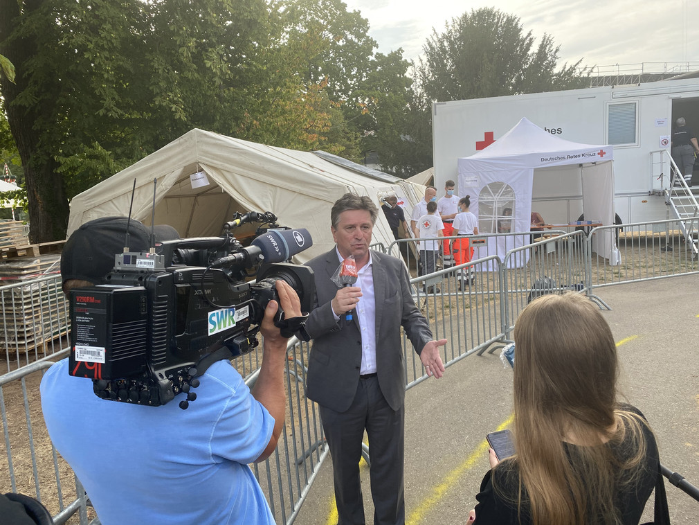 Gesundheitsminister Manne Lucha im Interview mit Reporterin, im Hintergrund die Coronavirus-Teststation am Stuttgarter Hauptbahnhof.