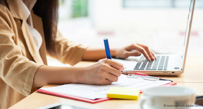 Junge Frau sitzt vor Laptop an einem Schreibtisch und macht sich Notizen.