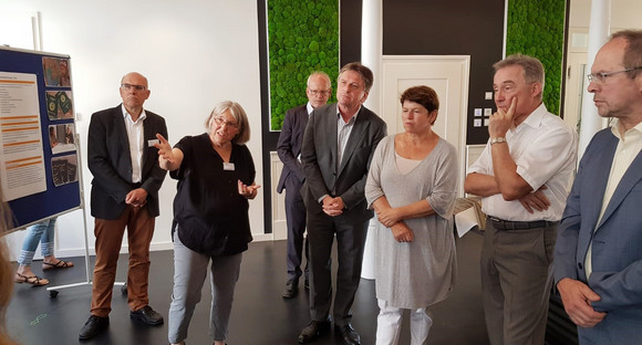 Minister Manne Lucha steht gemeinsam mit Silvia Maier-Lidle von der Ökumenische Fachstelle Asyl in Ludwigsburg vor Stellwänden einer Ausstellung