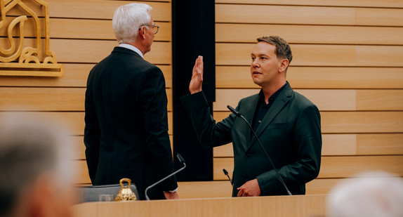 Oliver Hildenbrand steht auf der Tribüne des Landtags Baden-Württemberg mit erhobener Hand vor Landtagspräsident Thomas Strobl.