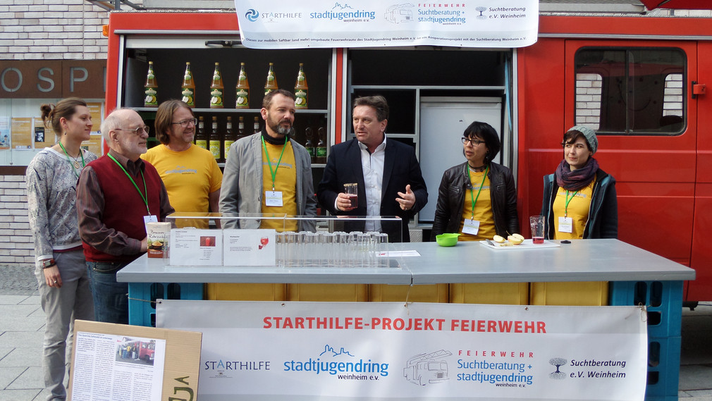 Gruppenfoto mit Minister Manne Lucha vor der „Feierwehr“, einem zur alkoholfreien Cocktailbar umgebauten alten Feuerehrauto der Starthilfekommune Weinheim.
